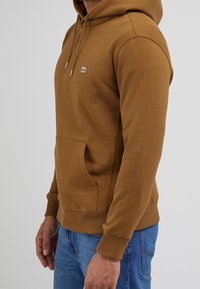 Ruskea puuvillainen huppari, jossa on etutasku, säädettävät kiristysnauhat, resorit hihansuissa ja pieni logo rinnassa.