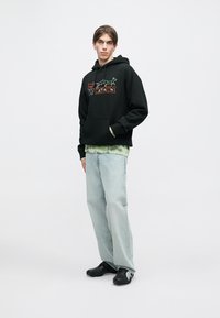 Μαύρο φούτερ με κουκούλα και γραφικό κείμενο σε πράσινο και πορτοκαλί, φορεμένο πάνω από ανοιχτό πράσινο πουκάμισο με σχέδιο tie-dye. Ανοιχτό μπλε τζιν και μαύρα παπούτσια.