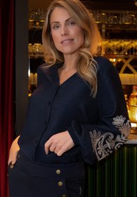 Marineblauwe blouse met knoopsluiting en lange mouwen, voorzien van uitgebreide gouden borduursels op de manchetten. Gecombineerd met een donkere rok die wordt geaccentueerd met gouden knopen.