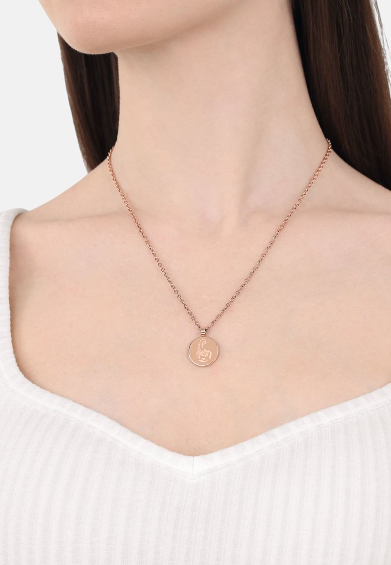 Halsband i roséguld med en graverad design, med en kedja och en cirkulär berlock, bärs mot en vit, ribbad topp.