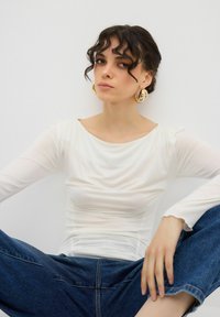 Vit långärmad topp med rynkad textur och rund halsringning, ihopparad med blå högmidjade jeansbyxor. Guldörhängen i form av ringar bärs.