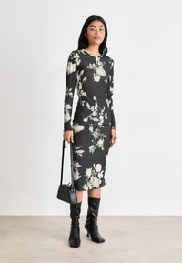 Blumengemustertes, langärmliges Kleid in Schwarz und Creme, figurbetonte Form, kombiniert mit knielangen schwarzen Stiefeln und einer kleinen Handtasche.