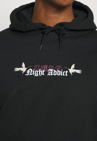 Sudadera negra con el texto "Night Addict" y dos ilustraciones de grúas, capucha con cordones y textura de tela suave.