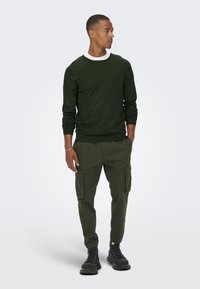 Donkergroen sweatshirt met ronde hals en lange mouwen, gecombineerd met bijpassende cargobroek met zijzakken. Zwarte veterschoenen maken de outfit compleet.