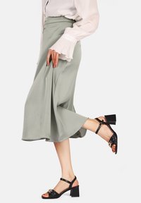 Gonna midi verde chiaro in tessuto morbido, abbinata a sandali neri con tacco a blocco e cinturino decorativo. Maniche a balze della blusa visibili.