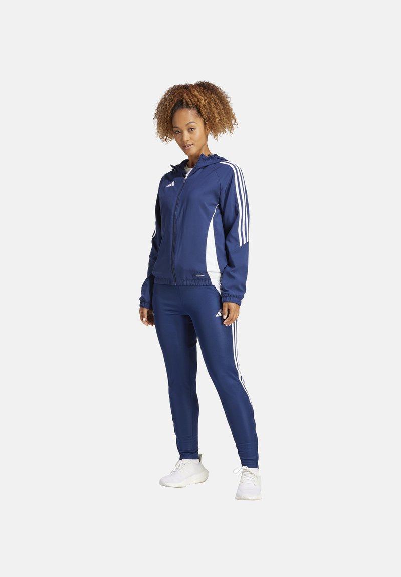 Tuta navy realizzato in tessuto liscio, con dettagli a strisce bianche su maniche e gambe, dotato di cappuccio con zip e design aderente.