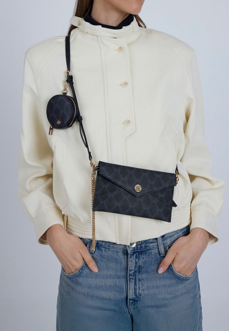 Černá prošívaná kožená kabelka crossbody se zlatým řetízkem, doplněná malou kulatou pouch na mince. Modelka má na sobě krémovou bundu a džínové kalhoty.