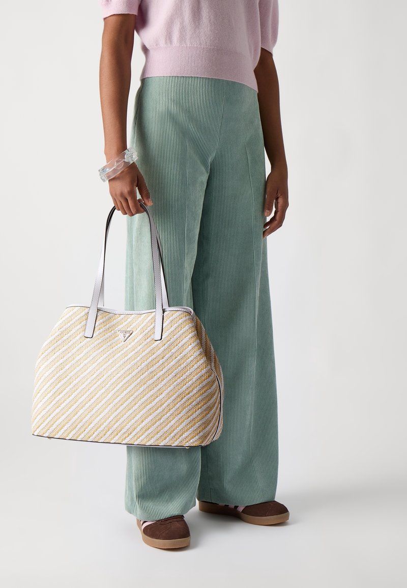 Personne portant un pantalon vert à jambes larges et un pull rose tenant un grand sac à main beige et blanc rayé avec des chaussures marron.