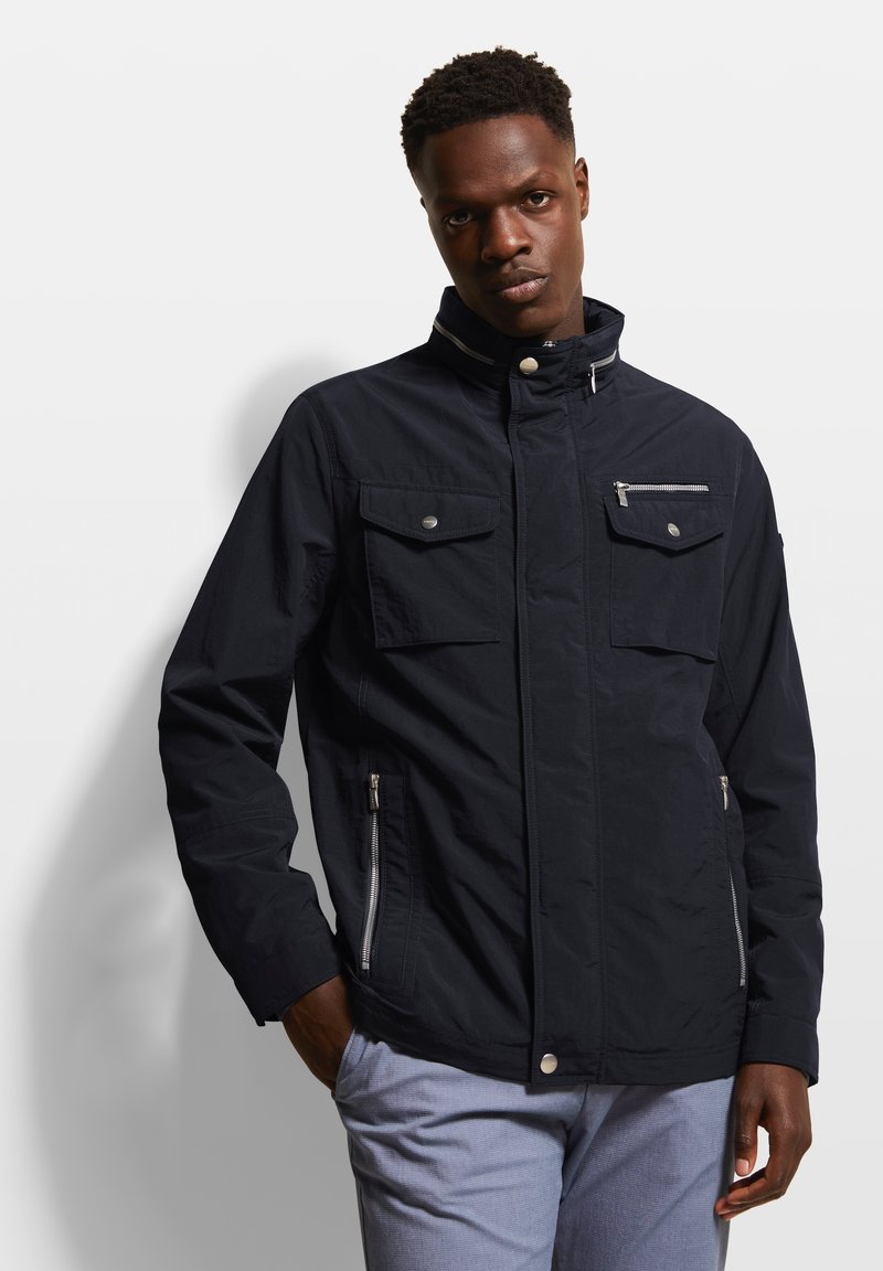 Marineblaue Zip-Jacke mit vier Taschen, Stehkragen und silbernen Hardwaredetails; aus einem leichten Material mit glatter Oberfläche hergestellt.