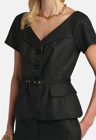 Frau trägt ein eng anliegendes schwarzes Kurzarm-Blazer mit Knöpfen, Gürtel und Pattentaschen, kombiniert mit einer goldenen Kettenhalskette und einem Armband.