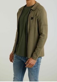 Homme portant une veste zippée vert olive sur un t-shirt vert foncé et un jean bleu clair déchiré, debout devant un fond clair uni.