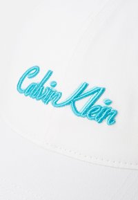 Căciulă de baseball albă cu text brodat în turcoaz "Calvin Klein". Prezintă un design structurat, material texturat și o vizieră curbată.