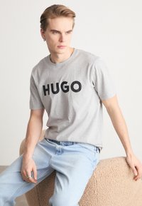 Pelēka kokvilnas T-kreklu ar uzrakstu "HUGO" treknā melnā fontā, savienots ar gaiši ziliem džinsiem, sēž uz teksturēta bēša otomā.