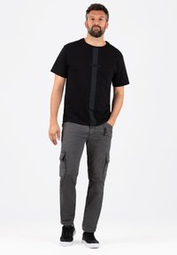 Schwarzes Rundhals-T-Shirt mit einem vertikalen dunklen Streifen und dem Schriftzug "TIMEZONE", kombiniert mit grauen Cargo-Hosen mit Seitentaschen. Schwarze Sneakers.