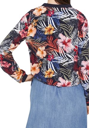 Femme portant un haut à manches longues bleu marine avec un imprimé floral tropical coloré et une jupe en denim bleu clair, la main reposant sur la hanche.
