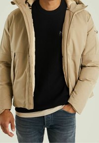 Beżowa ocieplana kurtka z kapturem, o gładkiej fakturze, z przednim zamkiem błyskawicznym i kieszeniami, noszona na czarnym swetrze dzianym i dżinsach.