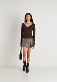 Μακρυμάνικη καφέ μπλούζα με V-λαιμόκοψη, συνδυασμένη με μια πλισέ γκρι καρό φούστα. Το μοντέλο κρατάει μια μικρή μαύρη τσάντα και φοράει μαύρες μπότες μέχρι τον αστράγαλο.