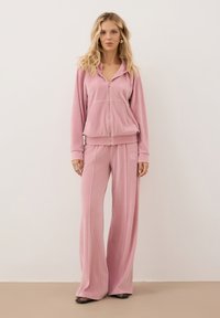 Tweedelig loungepak in zacht roze velours. Bevat een hoodie met rits en logo, en brede broekspijpen. Beide hebben een gladde textuur en een relaxte pasvorm.