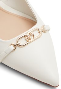 Chaussure plate blanche à bout pointu en cuir, avec un accent en chaîne dorée et un logo circulaire. Texture lisse et design minimaliste.