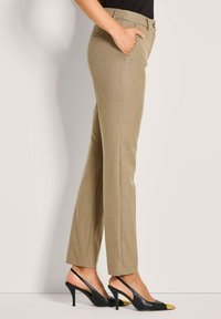 Vrouw in beige gestructureerde rechte broek met zijzakken en zwarte slingback hakken met gouden teenkapjes, staande in zijaanzicht.