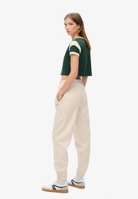 Chemise verte courte avec des manches crème, assortie à un pantalon de survêtement beige clair avec des poches latérales. Modèle portant des baskets blanches avec des accents noirs.