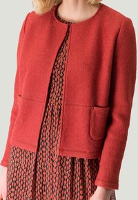 Červená texturovaná bunda s kulatým výstřihem, předními kapsami a rovného střihu, nošená přes vzorované šaty s kruhovými motivy v tmavých tónech.