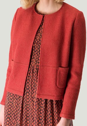 Veste texturée rouge avec un col rond, des poches avant et une coupe droite, portée sur une robe à motifs ondulés dans des tons sombres.