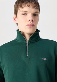 Grønn, langermet, halv-lynlåspullover med strukturert stoff. Har en liten brodert logo på brystet og en metallynlås.