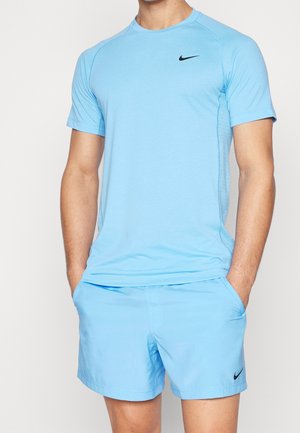 Man draagt lichtblauw Nike sport-t-shirt en korte broek, met handen in de zakken, staand tegen een effen achtergrond.