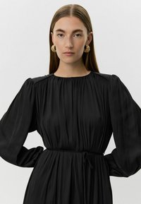 Juoda plisėta suknelė su ilgomis pūstomis rankovėmis, siaurėjančiu liemeniu ir apvaliu kaklu. Auksinės išsiskiriančios auskarų poros papildo aprangą.