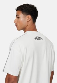 T-shirt blanc à manches courtes en coton. Présente un logo de marque discret en noir dans le dos et des coutures contrastantes le long des coutures d'épaule.