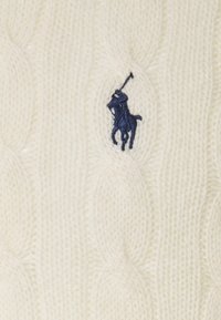 Stickad kabeltröja i kräm med ett strukturerat mönster, med en marinblå broderad polo-logotyp på framsidan. Mjukt ullmaterial.