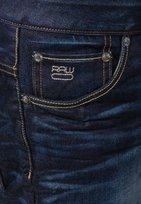 Mörkblå denimjeans med en blekt textur, med synliga sömmar, en framficka med knapp och en logotyp broderad på fickan.