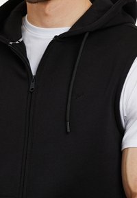 Sweat à capuche noir sans manches avec fermeture éclair, poches avant et cordon de serrage réglable. Présente un petit détail de logo sur la poitrine.