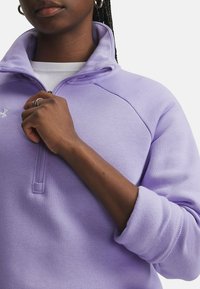 Oseba z nataknjeno pričesko nosi svetlo vijolično zip-up kapuco in drži ovratnik z eno roko pred enotno ozadjem.