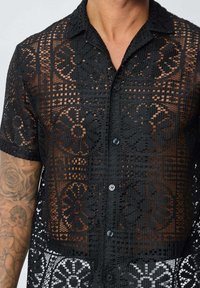 Chemise noire transparente à boutons présentant des motifs de dentelle complexes, des manches courtes et un col traditionnel, avec des motifs floraux détaillés.