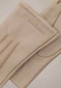 Beżowe skórzane rękawiczki z dekoracyjnym przeszyciem, z wytłoczonym logo na materiale. Gładka tekstura i dopasowany fason.