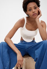 Hvid ribstrikket croptop med brede stropper, kombineret med løse blå denimbukser. Model siddende på en træoverflade og ser direkte ind i kameraet.