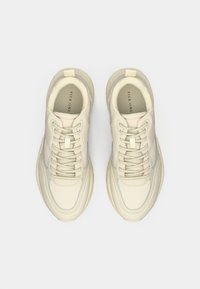 Sneakers krémovej farby s textúrovanou kožou, hrubou podrážkou, stehovaním v rovnakej farbe a jemným modrým akcentom pozdĺž bočnej strany. Štandardné šnurovanie.