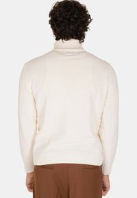 Maglione a collo alto crema con una texture a coste, caratterizzato da maniche lunghe e un orlo aderente, abbinato a pantaloni marroni. Vista posteriore mostrata.