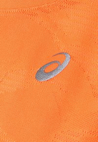 Světlá oranžová látka s texturovaným povrchem. Obsahuje stříbrné logo, které je výrazně zobrazeno, což naznačuje identifikaci značky.