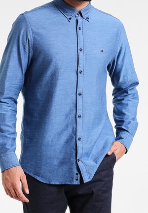 Mężczyzna w niebieskiej koszuli z długim rękawem, zapinanej na guziki, z zapiętym kołnierzykiem, z ręką w kieszeni, stojący na białym tle.