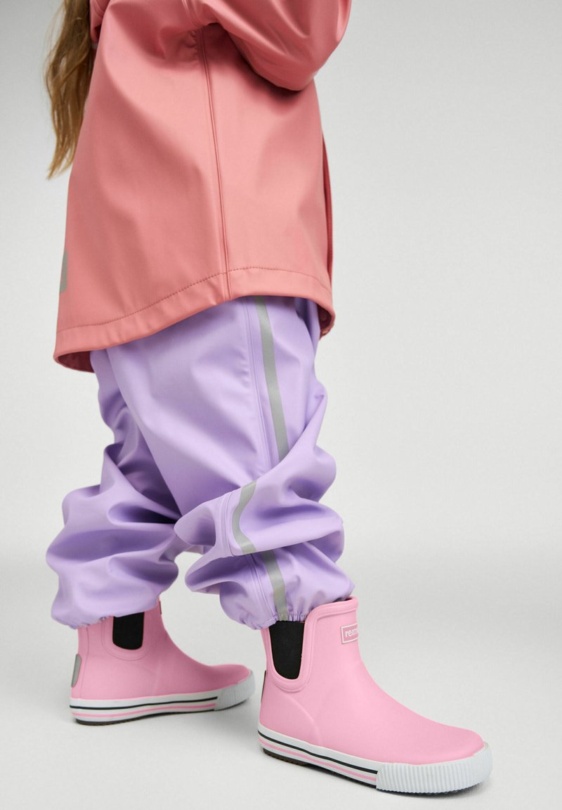 Enfant portant des bottes de pluie roses, un pantalon imperméable lavande avec des bandes réfléchissantes, et une veste de pluie rose, debout sur un fond clair.