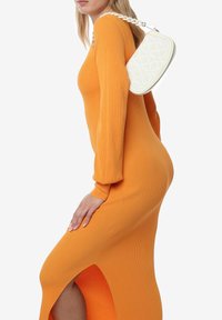 Geribbelde langetermijndress in fel oranje, met een zijsplit. Een witte, gewatteerde handtas met een gevlochten schouderband wordt op de schouder gedragen.
