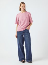 T-shirt en coton rose avec des détails en œillets sur les manches, associé à un jean en denim foncé à jambes larges. Le modèle se tient avec une main dans la poche.