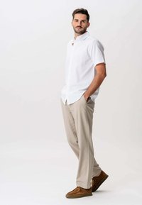 Hvid kortærmet skjorte med knapper, beige dressbukser og brune mokkasiner. Model står med hænderne i lommerne foran en ensfarvet baggrund.