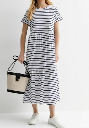 Femme portant une robe midi blanche à rayures horizontales noires, des baskets blanches, et tenant un sac à main beige et noir avec une bandoulière.