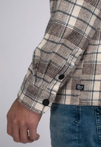 Flanellhemd im Karomuster mit Buffalostreifen in Creme, Braun und Blau. Inklusive schwarzer Knöpfe an den Manschetten und einem Logoschild am Saum.