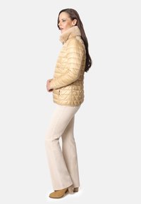Chaqueta acolchada beige con cuello de piel sintética, que cuenta con bolsillos laterales y cierre de cremallera, combinada con pantalones de campana de color claro y botas marrones.