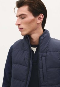 Gilet imbottito blu navy con colletto alto e chiusura con zip, caratterizzato da una texture trapuntata e una piccola tasca esterna sul petto.